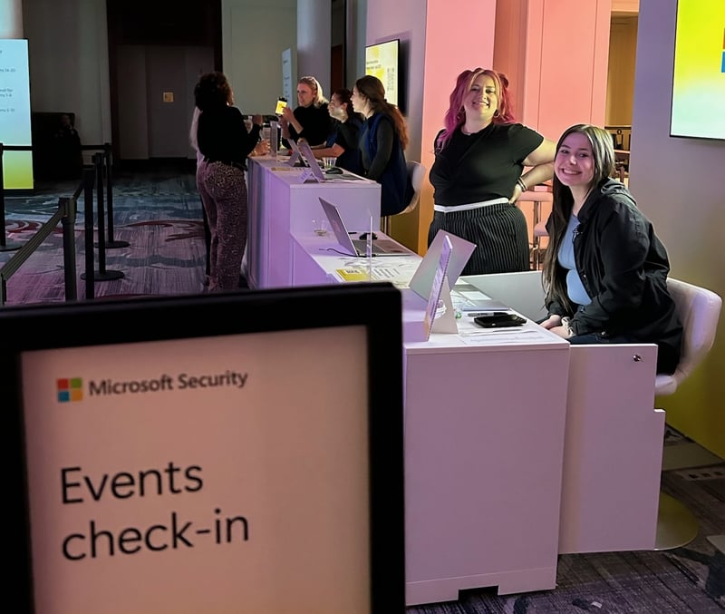 Two women are behind a check in desk. Both are smiling towards the camera.