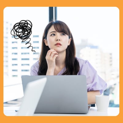 A female Asian office worker, sits behind a laptop. She looks perplexed, a scribbled cloud of jumbled thoughts is drawn by the side of her head