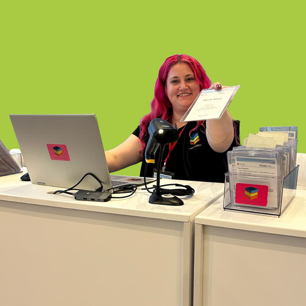 A young woman with red hair sits behind a desk, she is holding out an event lanyard and smiling.