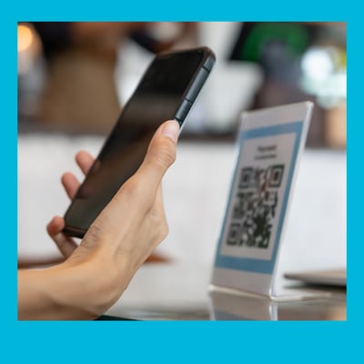 A photo with a blue border of a mobile phone pointed towards a QR code on a desk