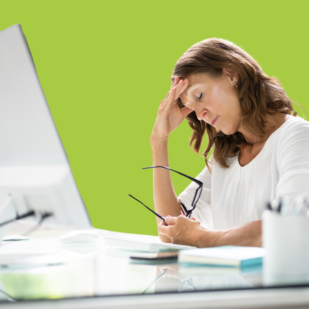 A white woman sits at a desk in front of a computer, she holds her glasses in one hand and rubs her temples with the other.