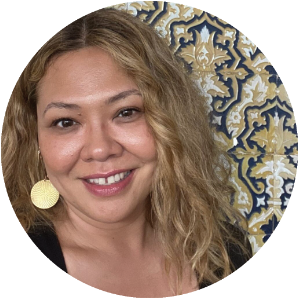 A headshot of an Asian woman with long wavy blonde hair, smiles warmly. She stands against a blue, brown and white patterned background. She is wearing a black top and large flat circled earrings.