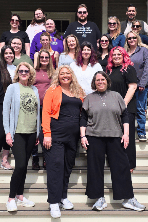 Sixteen members of the EventBuilder team stand on a set of steps, at the front the management trio of Robin, Renee and Robin, are smiling warmly and looking relaxed.