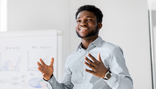 A Black man in a light blue shirt smiles and presents in front of a standing white board with graphs and charts on it