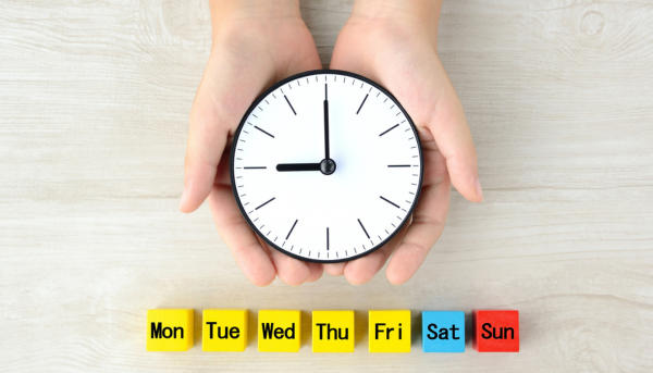 A pair of female hands hold a clock face, with yellow blue and red blocks underneath with days of the week on them