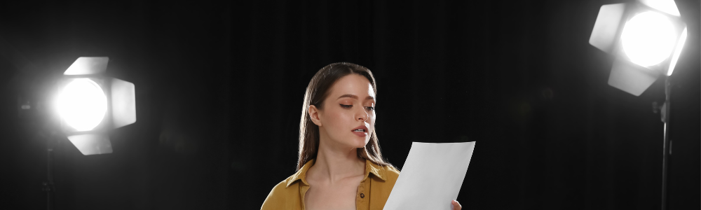 A woman reads from a script there are stage lights in the background to her left and right