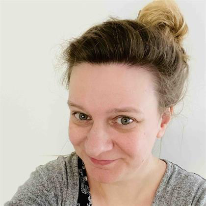 A woman smiles bashfully at the camera, her hair is in a messy bun.